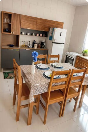 Un comedor con una mesa dispuesta para cuatro, sillas de madera, un jarrón con flores y una cocina al fondo.