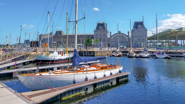 Ein Yachthafen mit vertäuten Booten, ein großes Gebäude mit markanter Architektur im Hintergrund und eine Fußgängerbrücke über dem Wasser.