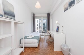 Ein modernes, helles Schlafzimmer mit Bett, Schreibtisch und Stuhl, einem Balkon mit Aussicht und Kunstwerken an den Wänden.