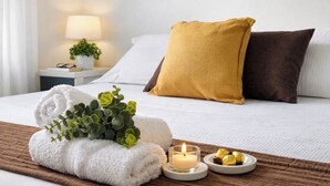 Una cama bien hecha con almohadas blancas y marrones, una toalla blanca y una planta pequeña sobre un tapete marrón. Hay una vela encendida y bombones en un plato.