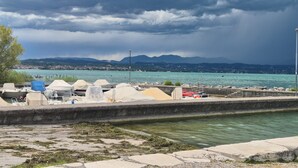 Vista al agua con barcos cubiertos por lonas, un camino de piedra y una cordillera distante bajo un cielo nublado.