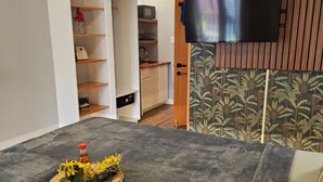 Uma sala de estar moderna com uma televisão de tela plana fixada em um painel de madeira, uma mesa de jantar com um centro decorativo e uma área de cozinha com armários de madeira e micro-ondas.