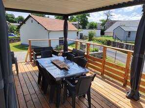 A covered outdoor deck with a table and chairs, a view of houses in the background, and a wooden railing. - Le Gite de Louisantin 6 people clim swimming pool (Lacapelle-del-Fraisse)
