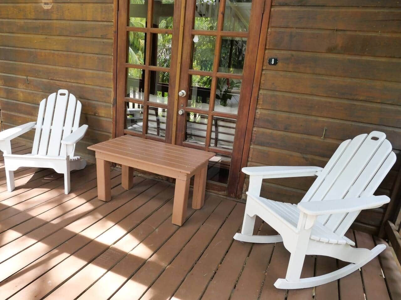 Een houten terras met twee witte Adirondack-stoelen en een kleine tafel.