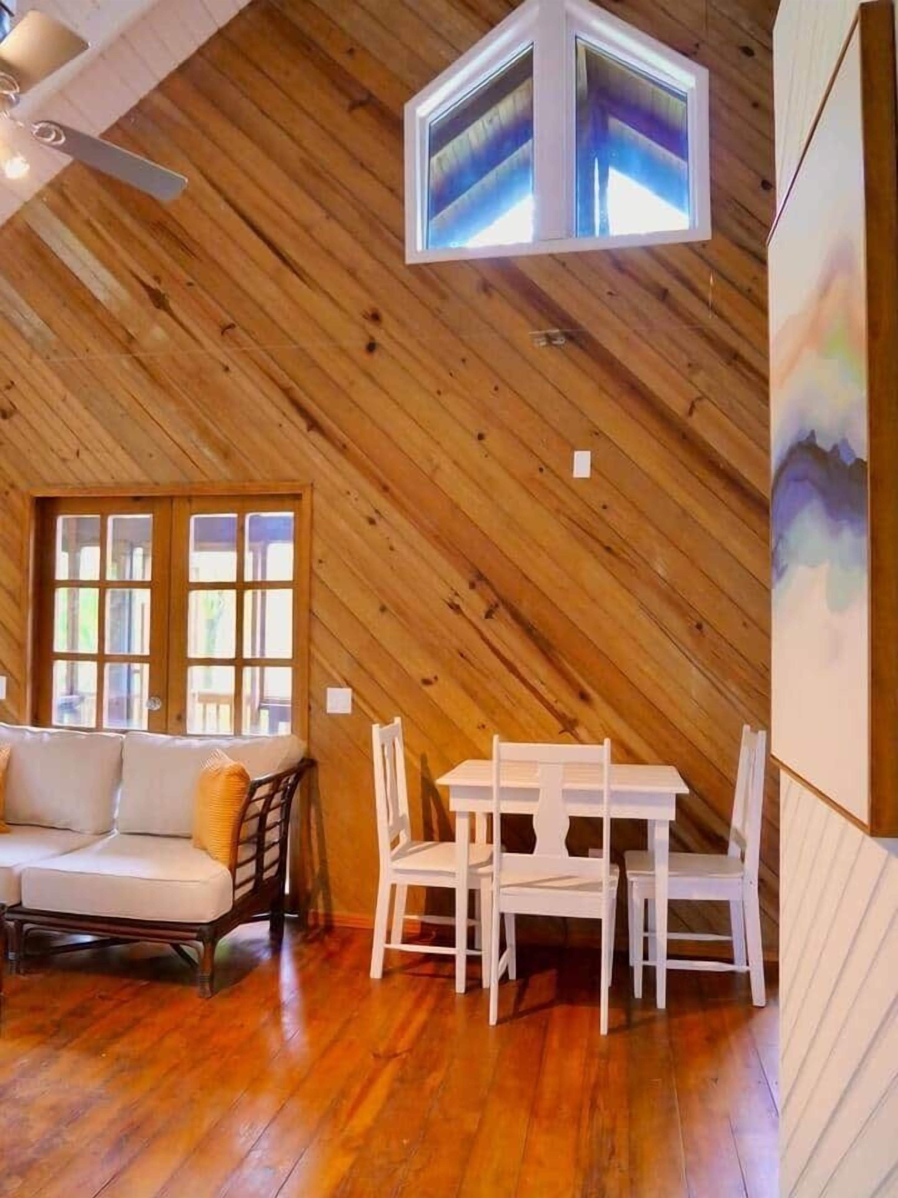 A room with a sloped wooden ceiling, a white sofa with yellow cushions, a white dining table with chairs, and a large window with multiple panes.