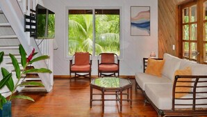 A room with a wooden floor, a white sofa, two red chairs, a glass coffee table, and a large window with a view of palm trees.