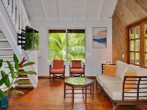 A room with a wooden floor, a white sofa, two red chairs, a glass coffee table, and a large window with a view of palm trees. - Sundancer 2 Br Home access pool beach dock (Sandy Bay)