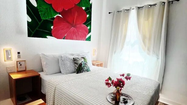 Une chambre avec un grand lit, une table de chevet, une lampe de chevet, un luminaire fixé au mur et une fenêtre avec des rideaux.