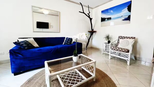 A living room with a blue sofa, a white coffee table, a white armchair, and a wall-mounted TV. - Dover Apt #3, Beach 5 mins, St Lawrence Gap (Oistins)