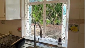 A kitchen with a stainless steel sink, a window with a metal grid, and a view of greenery outside. - Dover Apt #3, Beach 5 mins, St Lawrence Gap (Oistins)