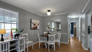 Un comedor con una mesa de madera y sillas blancas, una consola blanca con objetos decorativos, y un ventanal con persianas.