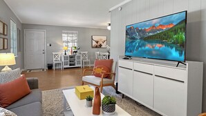 Una sala de estar moderna con un televisor de pantalla plana, un sofá gris, una mesa de centro con objetos decorativos y un comedor al fondo.