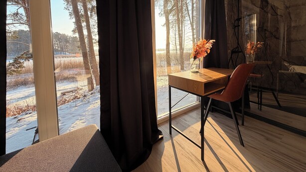 A modern room with a large window, a wooden table, a chair, and a vase with flowers. - Lakefront Cabin w/ Sauna & Hot Tub | Aiyana Retreat (Gauja)