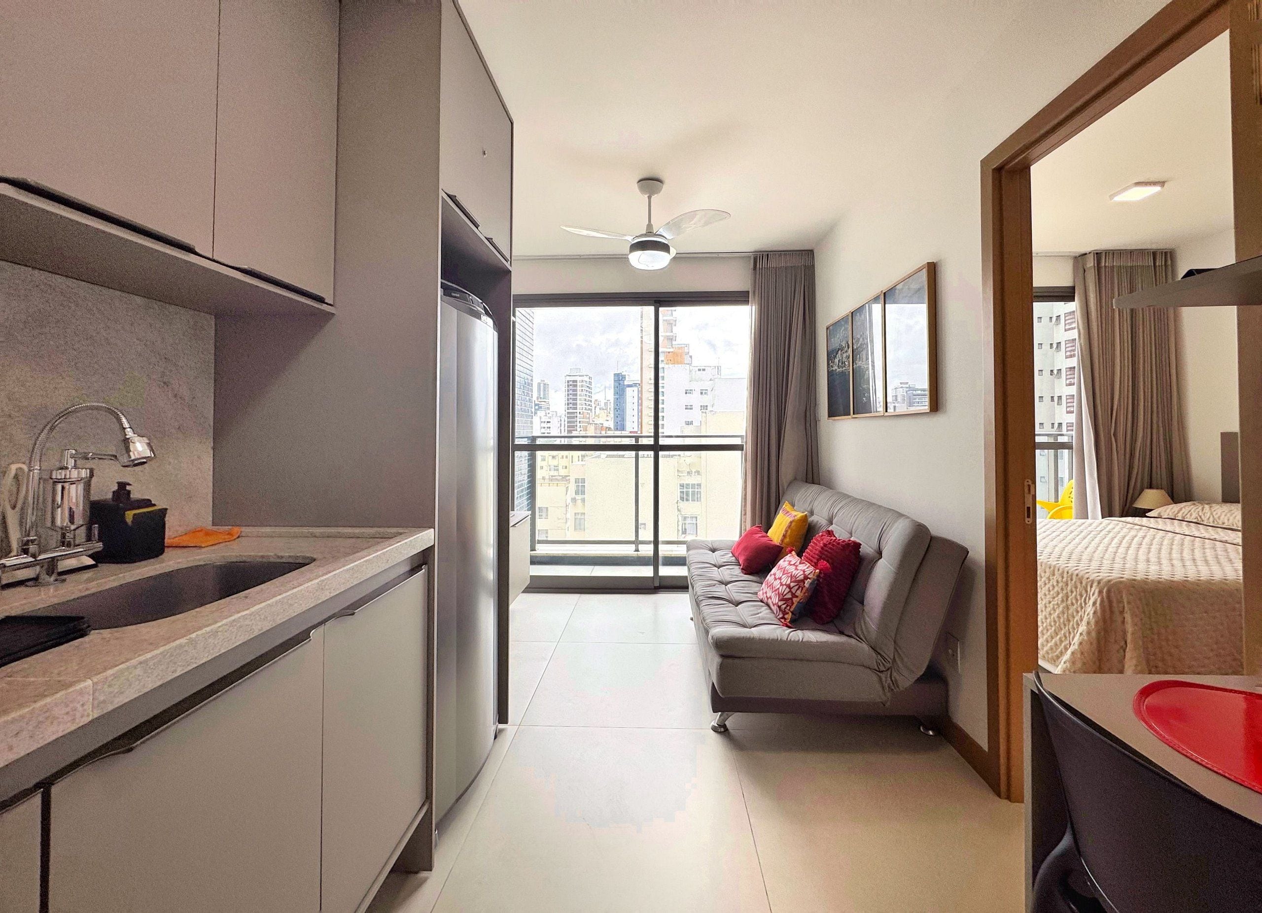 Un appartement moderne doté d’une kitchenette, d’un canapé gris agrémenté de coussins colorés, d’une grande fenêtre offrant une vue sur la ville et d’un ventilateur de plafond.