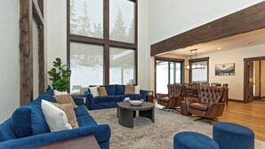 A spacious living area with a large window, a blue sofa set, a round coffee table, and a wooden ceiling. - Plum Creek Lodge • Expansive Mountain Views • 6BR • 5.5BA • Game Room (Cle Elum)