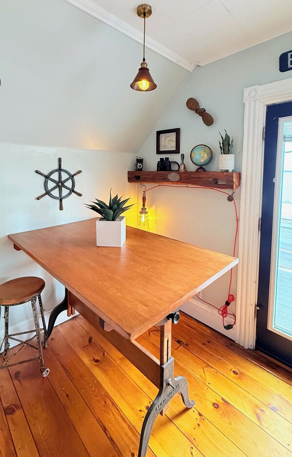 A wooden dining table with a plant, a ship's wheel, and a hanging light fixture in a room with wooden flooring and a door in the background. - Casco Bay Vista (Portland)