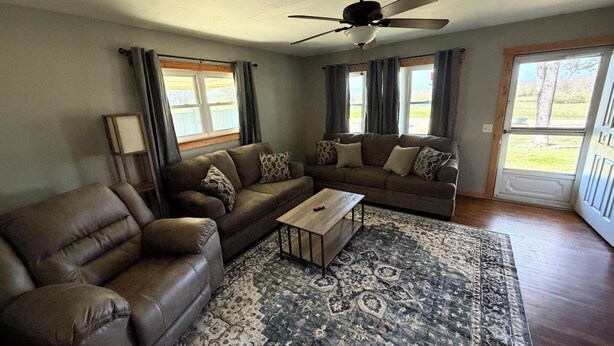 A living room with a sofa, armchair, coffee table, patterned rug, ceiling fan, and large windows with curtains. - Mountain-View 3BR Home w/ Firepit • Hatfield AR (Hatfield)