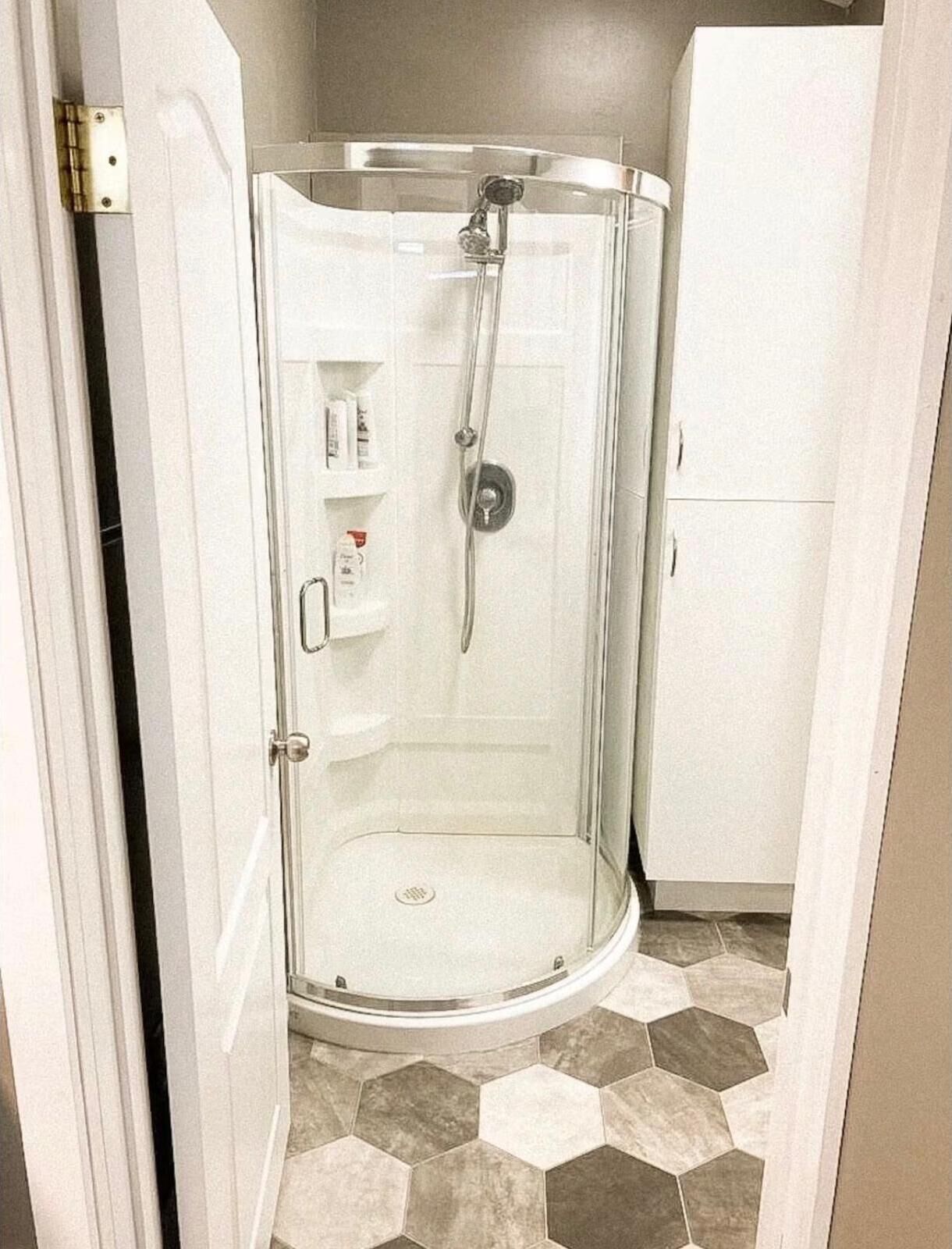 A glass-enclosed shower stall with a white door, a small shelf with toiletries, and a hexagonal tile floor.