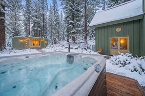 A hot tub surrounded by snow-covered trees, a wooden deck, and a cabin with a lit interior.