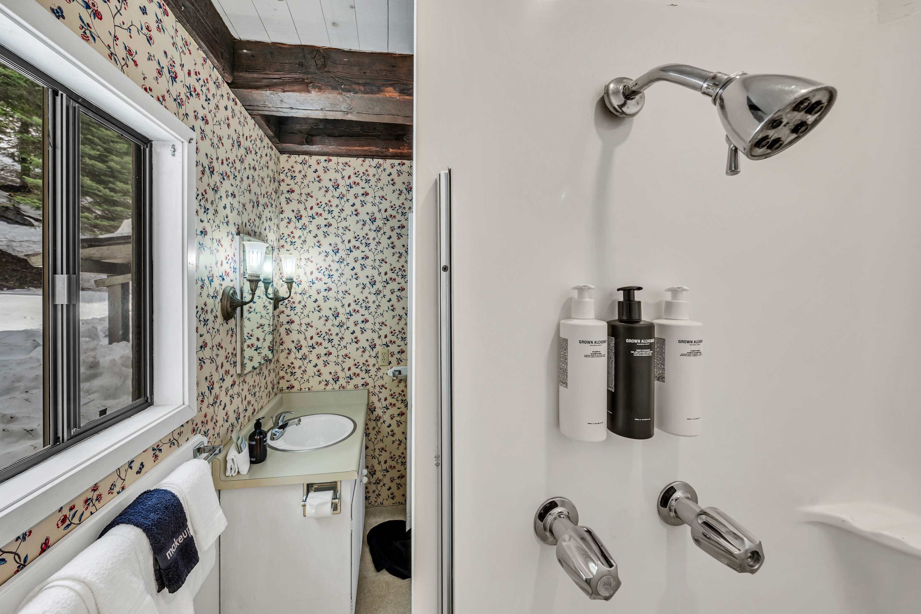 A bathroom with a floral wallpaper, a sink with a mirror, a towel rack, and a shower with a handheld showerhead.