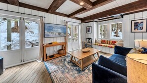 A cozy room with wooden beams, a large window with a view of snow-covered trees, a wooden table, a blue sofa, and a television on the wall. - Olympic Valley Escape - Riverside Views + Hot Tub (Tahoe City)