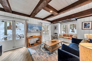 A cozy room with wooden beams, a large window with a view of snow-covered trees, a wooden table, a blue sofa, and a television on the wall. - Olympic Valley Escape - Riverside Views + Hot Tub (Tahoe City)