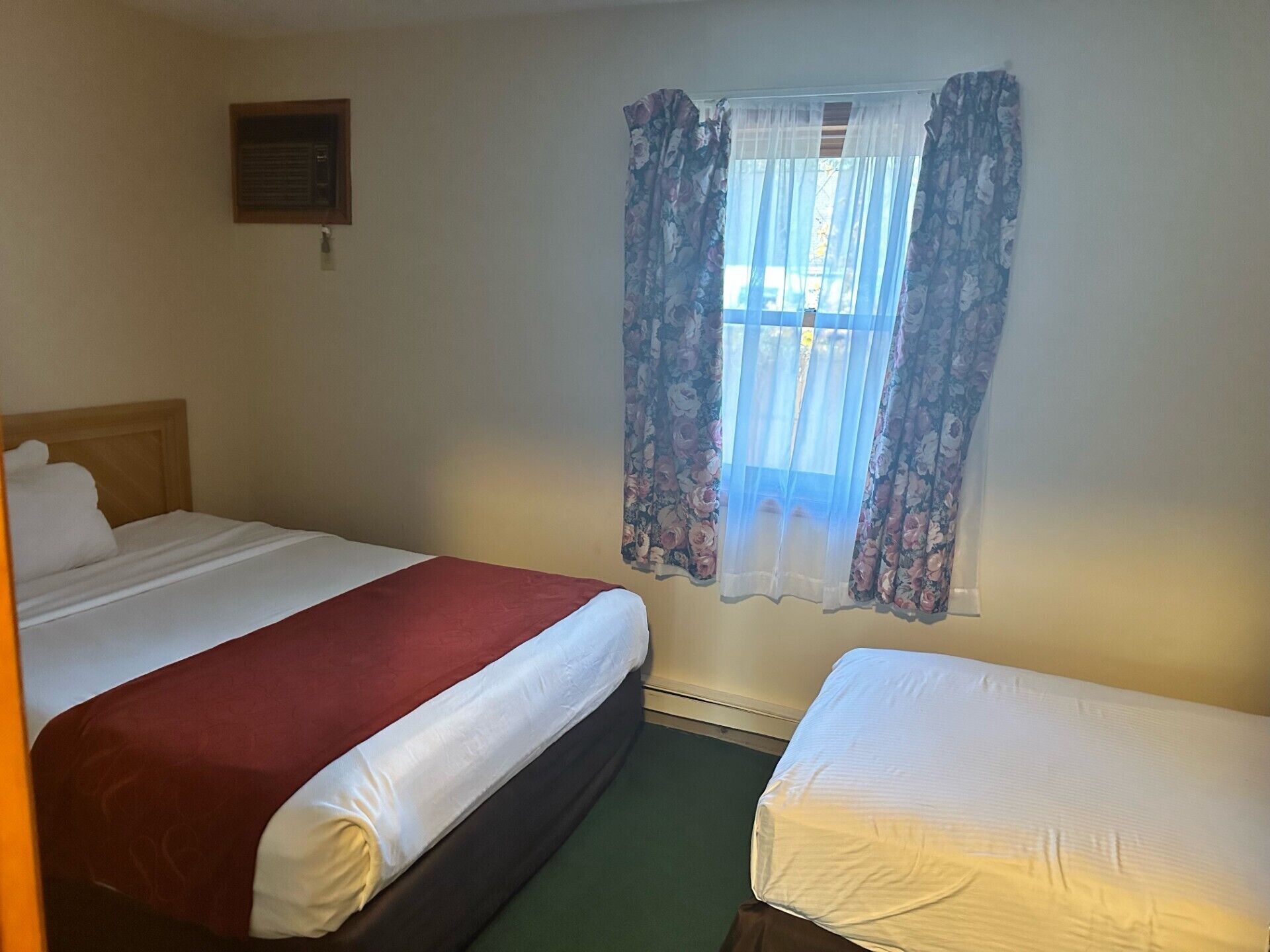 A hotel room with two beds, one with a red bedspread and the other with a white bedspread. There is a window with floral curtains, a small air conditioning unit on the wall, and a green carpet.