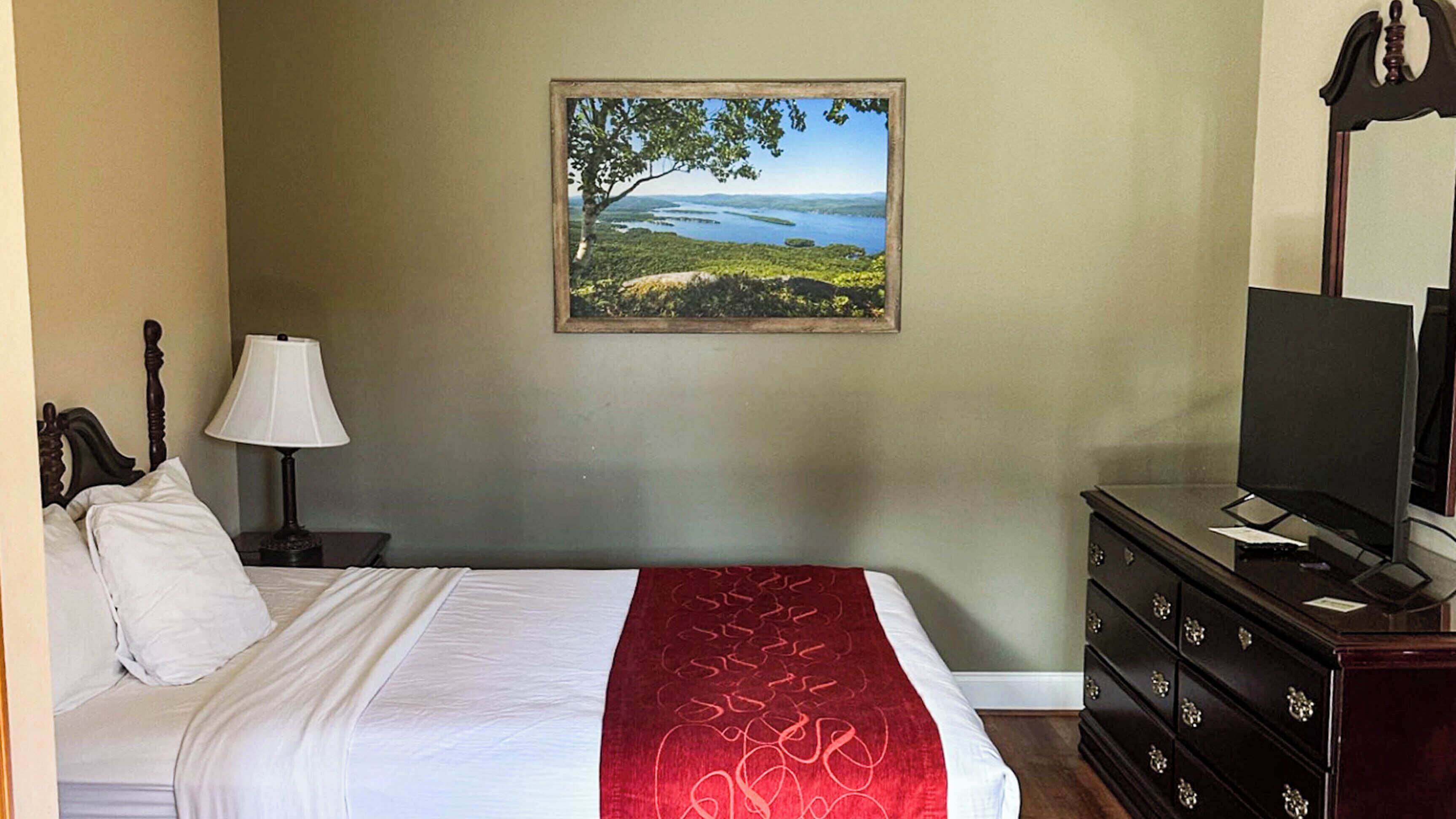A hotel room with a bed, a TV, a dresser, a lamp, and a painting of a landscape.