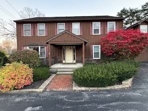 A two-story building with a front porch, a brick pathway leading to the entrance, and a well-maintained garden with shrubs and a flowering tree.