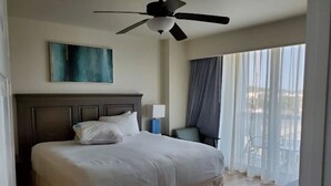 Een hotelkamer met een bed, een plafondventilator, een schilderij aan de muur, een raam met gordijnen en uitzicht op buiten.
