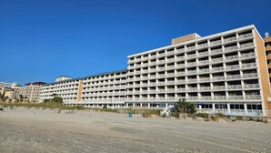 Een hotel met meerdere verdiepingen en veel balkons, een zandkleurige begane grond, en een helderblauwe lucht.