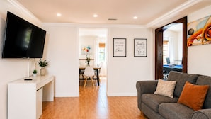 Una sala de estar con un televisor de pantalla plana, un sofá gris con cojines, un comedor con mesa y sillas, y dos citas inspiradoras enmarcadas en la pared.