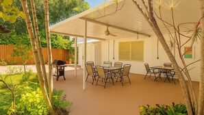 Un patio techado con mesa y sillas, una parrilla y vistas a un jardín con árboles y una valla.