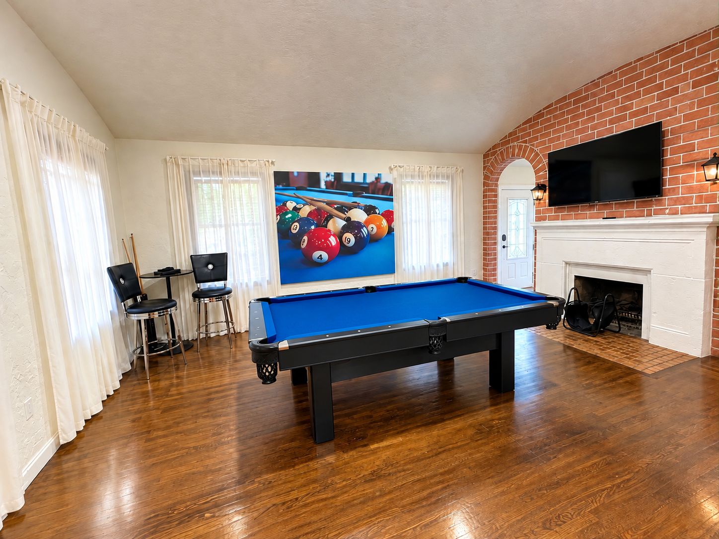 A pool table in a room with a brick fireplace, a television mounted above it, and a billiards cue rack.