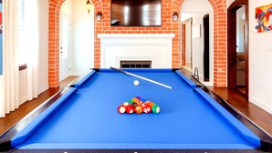 Una mesa de billar con superficie de fieltro azul, rodeada de paredes de ladrillo rojo, una chimenea blanca y un televisor de pantalla plana montado.
