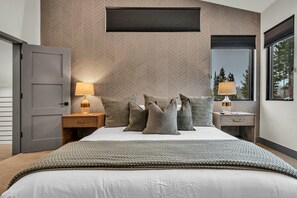 Un dormitorio moderno con una pared de diseño en patrón espiga, una cama grande con un plaid tejido, dos mesitas de noche con lámparas y una ventana con vistas a árboles.