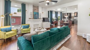 A spacious living area with a green sofa, yellow armchair, and a dining table with chairs. A fireplace and a television are also visible.