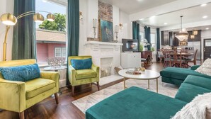 A modern living room with a fireplace, green and yellow armchairs, a dining area, and a kitchen in the background.