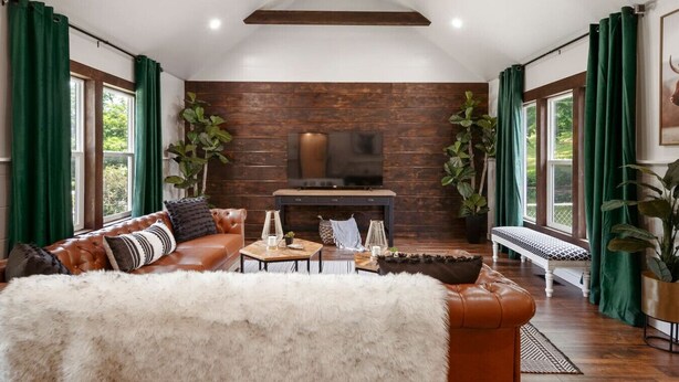 Een moderne woonkamer met een leren bank, een witte bontdeken, een open haard en grote ramen met groene gordijnen.