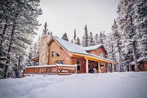 Une cabane en rondins avec une cheminée en pierre, une terrasse en bois et entourée d’arbres couverts de neige.