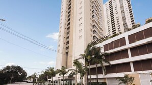 Un edificio residencial alto con varios balcones, una estructura de estacionamiento con área cubierta y palmeras en primer plano.