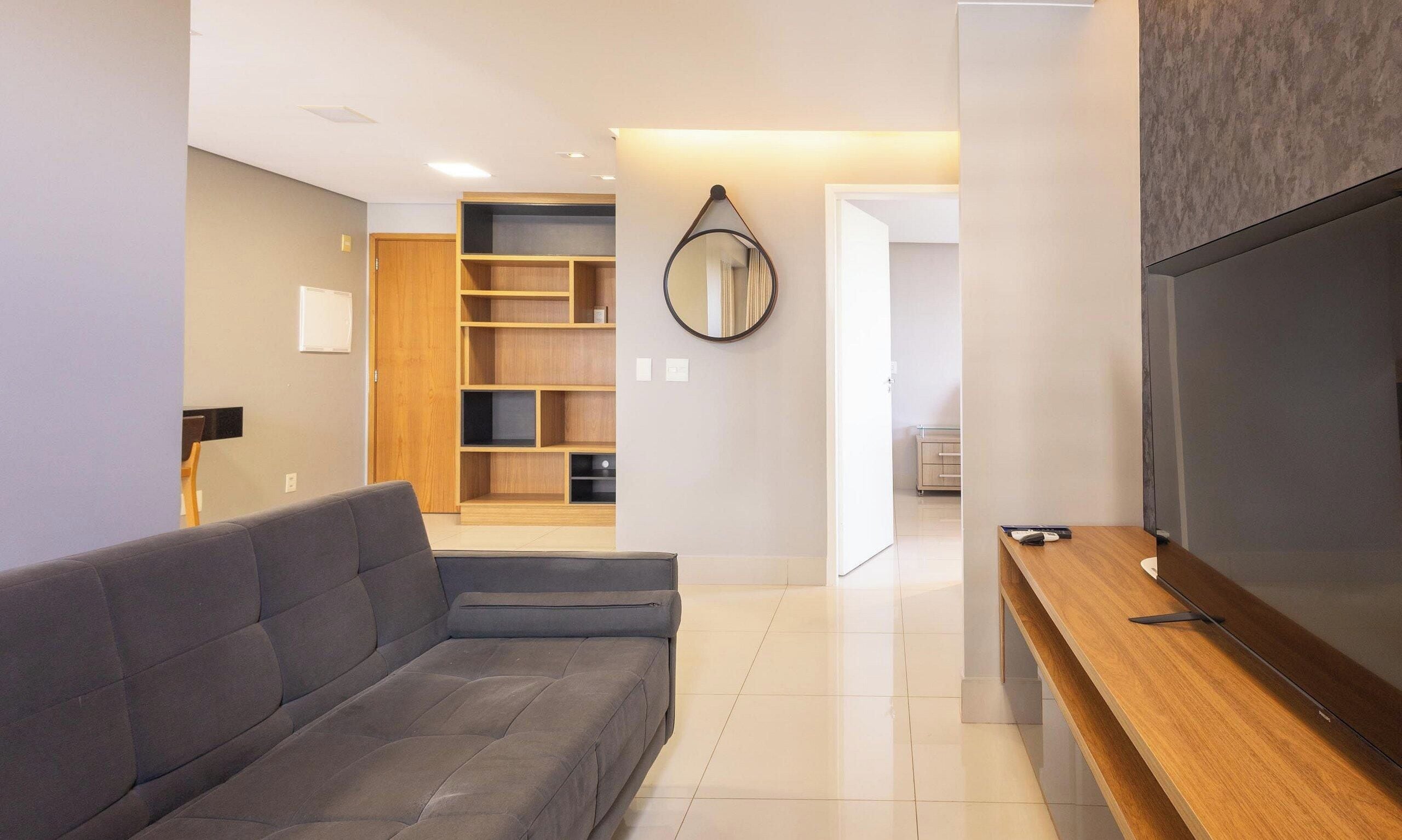 A modern living room with a grey sofa, wooden shelving, a mirror, and a TV on a wooden stand.