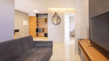 A modern living room with a grey sofa, wooden shelving, a mirror, and a TV on a wooden stand.