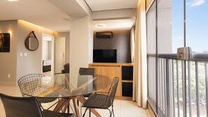 Un comedor moderno con una mesa y sillas de vidrio, un televisor montado en un panel de madera, y un balcón con vistas.