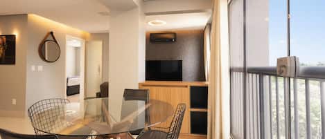 A modern dining area with a glass table and chairs, a TV mounted on a wooden panel, and a balcony with a view.