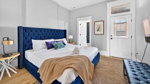 A bedroom with a tufted headboard, a bed with white and purple bedding, a bedside lamp, a wooden side table, a bench, a ceiling fan, and a door with a window. - 7Br Group Haven: Yard • Fire Pit • 3 Kitchen (St. Louis)