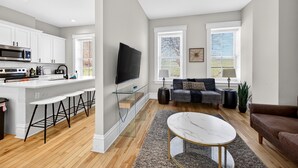 A modern kitchen with white cabinets, a large island with bar stools, a flat-screen TV, and a ceiling fan. - 7Br Group Haven: Yard • Fire Pit • 3 Kitchen (St. Louis)