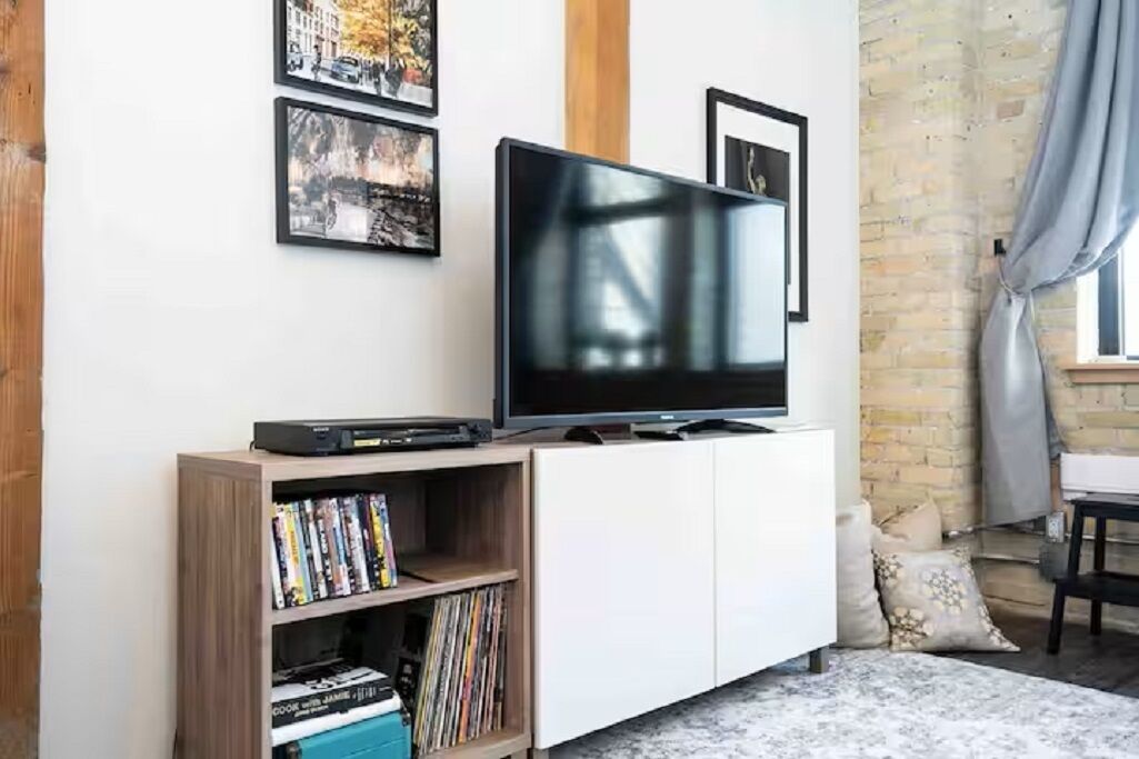 Un salon avec un téléviseur à écran plat posé sur une étagère en bois, un meuble blanc et un mur en briques.