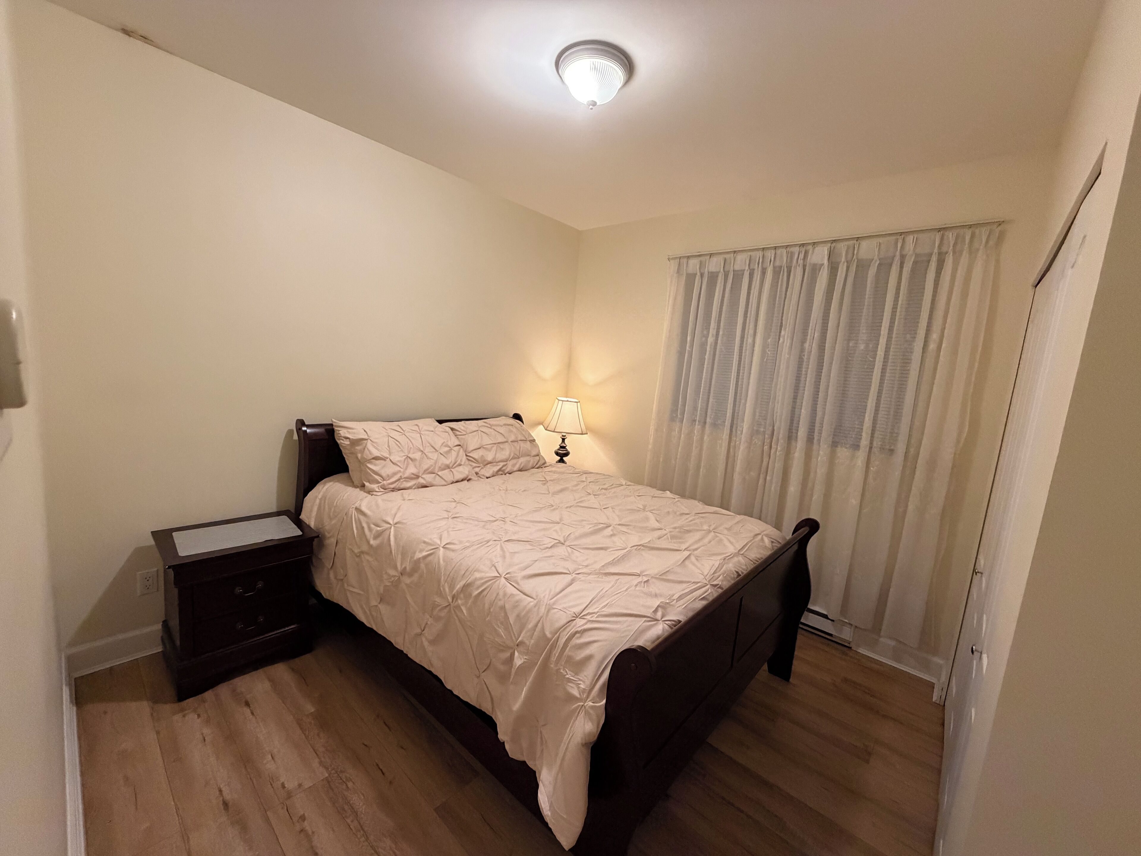 Et soveværelse med seng, et natbord, en lampe, et vindue med gardiner og en loftlampe.
