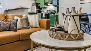 Una sala de estar con un sofá de cuero marrón, una mesa redonda y una cocina al fondo.